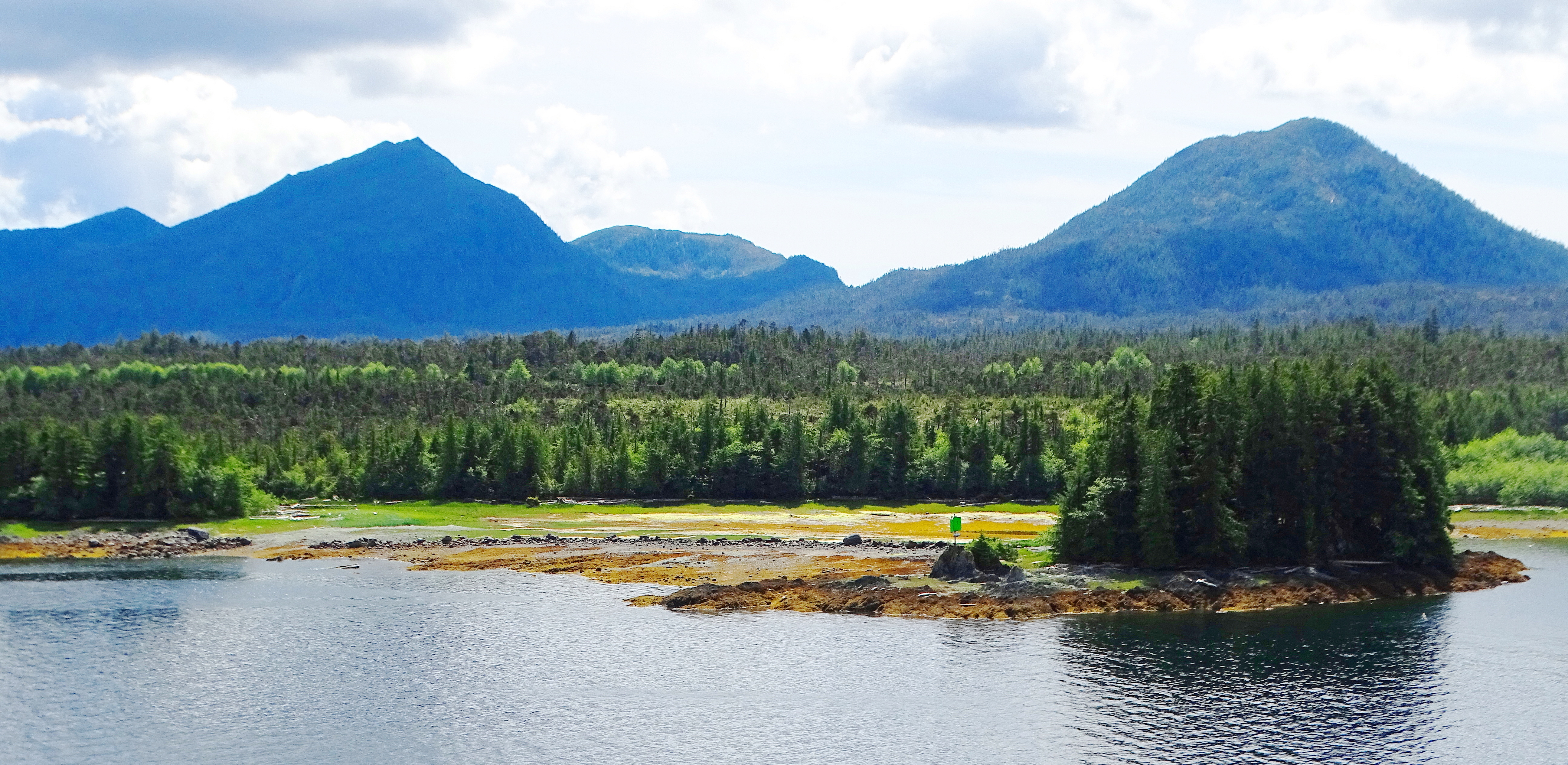 A scenic view of a tranquil body of water surrounded by lush greenery and rolling mountains in the background, featuring a small rocky island with trees and a green marker.