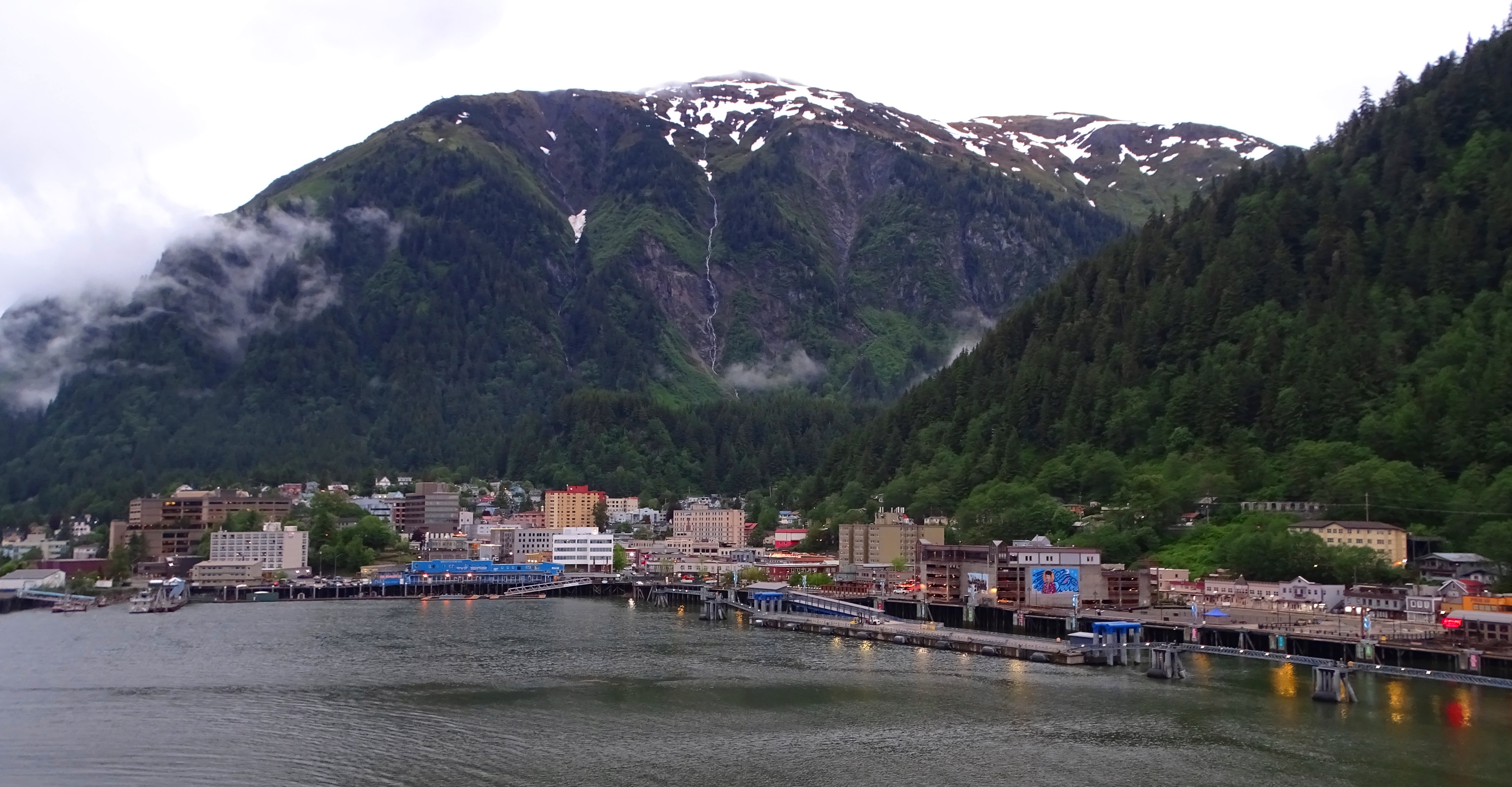 Scenic view of a coastal town surrounded by mountains, with buildings along the shore and misty peaks in the background.